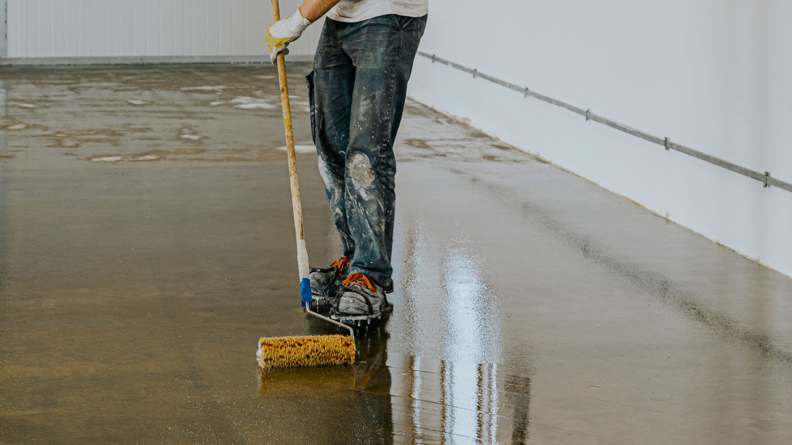 A person applying floor paint with a roller on a concrete surface.